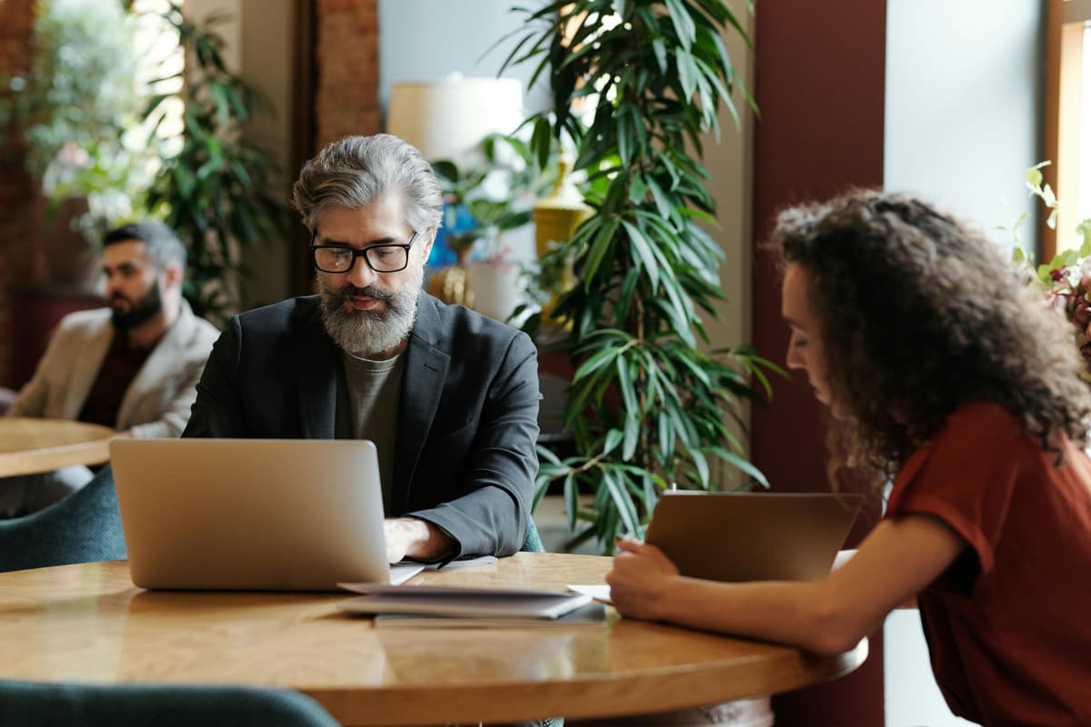 A professional man with a grey beard and glasses working on a laptop at a wooden table in a modern, plant-filled office space with a colleague.