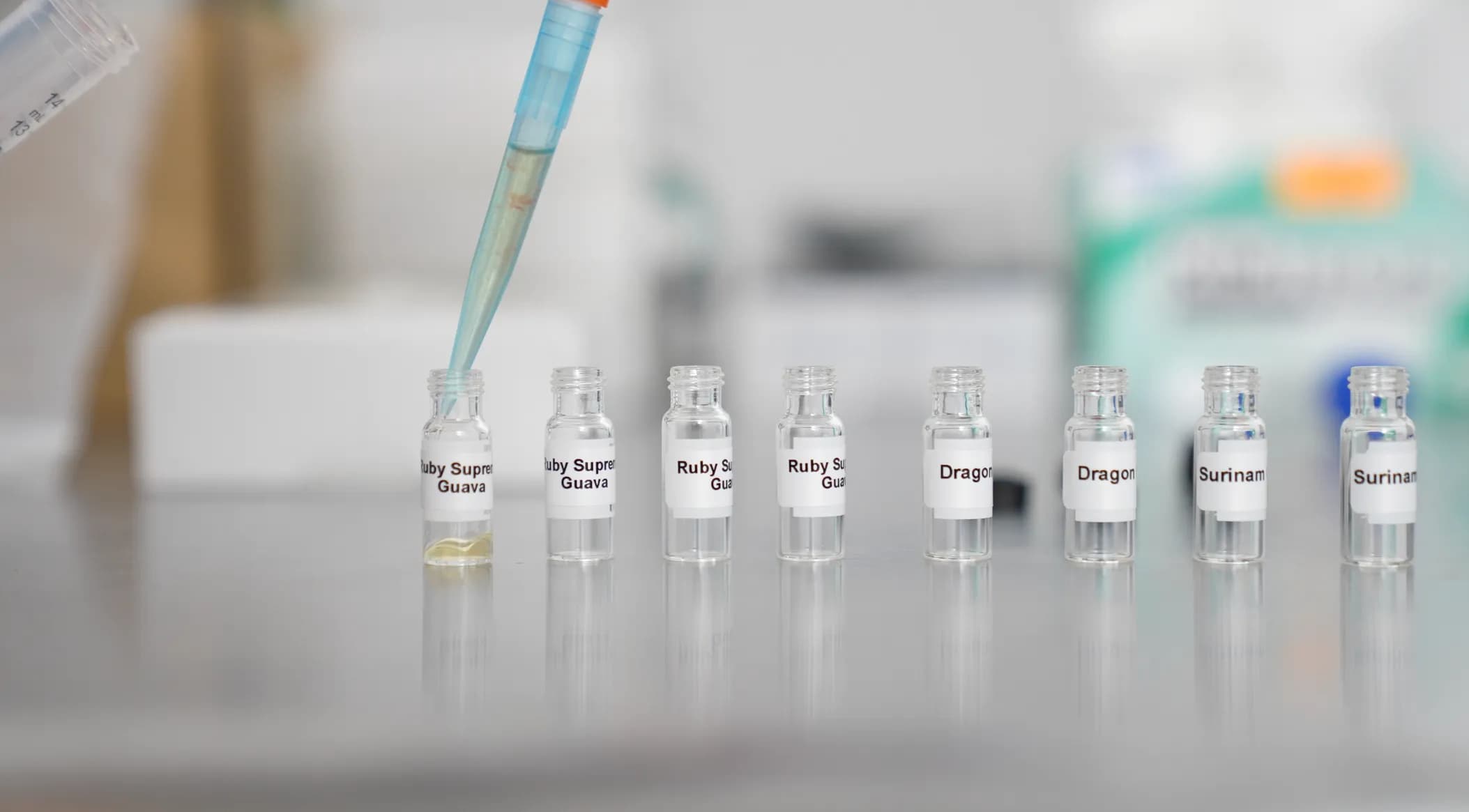 A row of glass vials containing terpene samples labeled 'Ruby Supreme Guava', 'Dragon', and 'Surinam'. A lab pipette is shown precisely dispensing a golden-colored liquid into one of the vials in a sterile laboratory environment