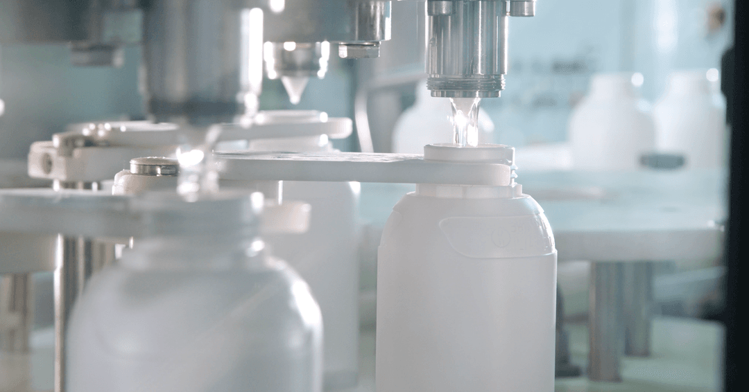An industrial automated bottling line in a sterile environment. A stainless steel nozzle precisely fills a white, opaque plastic bottle with a clear liquid. Several other similar bottles are visible on the moving conveyor system, held in place by white mechanical guides under a bright, clinical light.