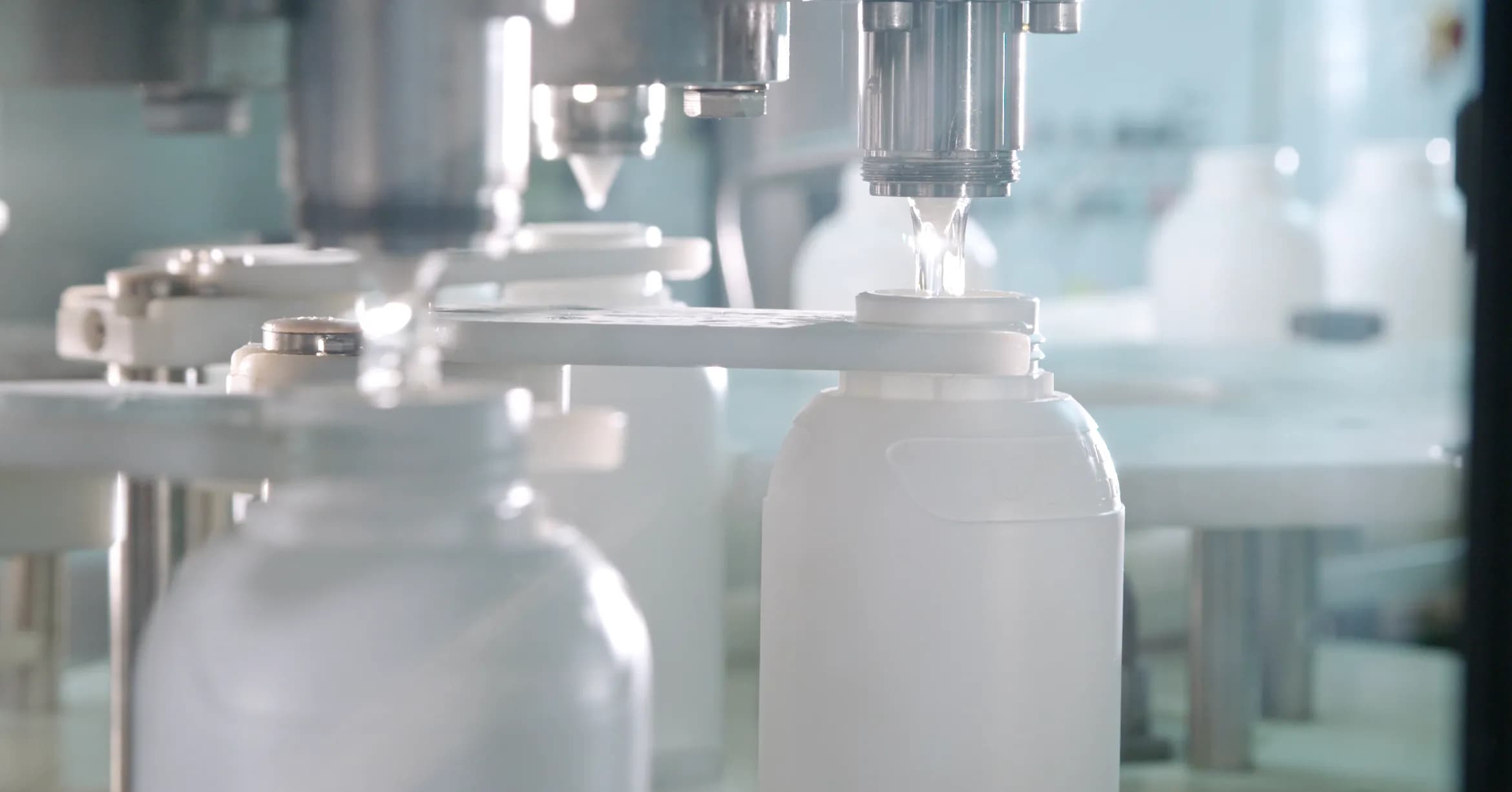 An industrial automated bottling line in a sterile environment. A stainless steel nozzle precisely fills a white, opaque plastic bottle with a clear liquid. Several other similar bottles are visible on the moving conveyor system, held in place by white mechanical guides under a bright, clinical light.