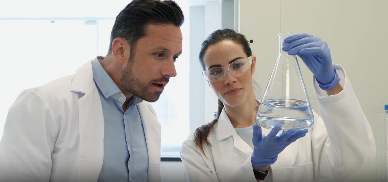 Two scientists in white lab coats and safety glasses closely examining a clear liquid inside a glass Erlenmeyer flask in a laboratory setting.